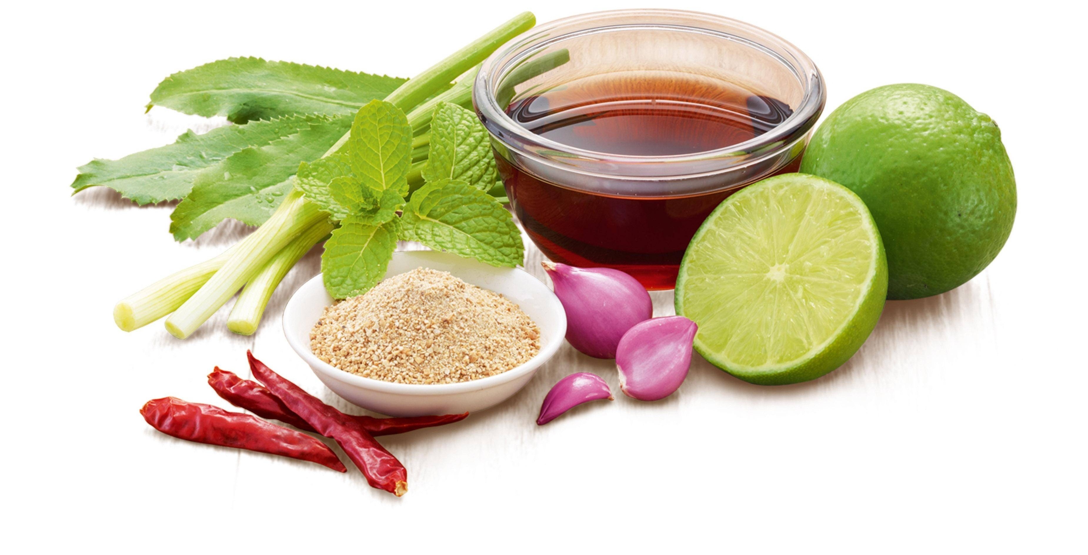 Ingredientes frescos como limas, chiles rojos, chalotas, hierba de limón, hojas de menta, un cuenco de salsa oscura y arroz tostado sobre una superficie rústica, en tonos verdes y terrosos