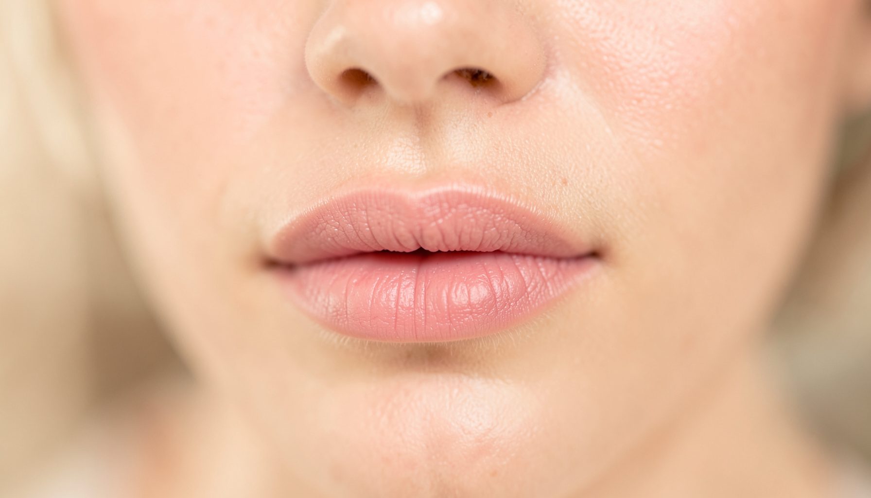 Primer plano de labios suaves con un ligero tono rosado, iluminación cálida y difusa, fondo desenfocado en tonos crema y beige, atmósfera serena y cuidada.
